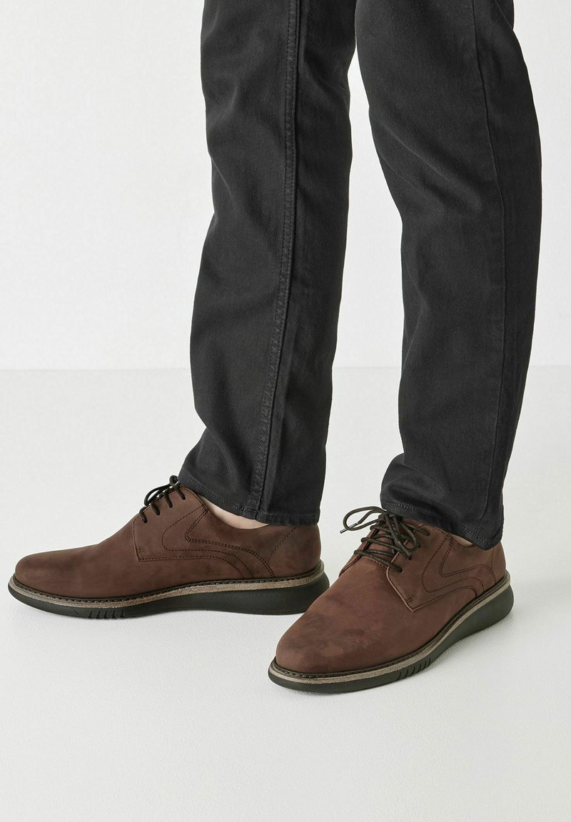 Bruine leren veterschoenen met een ronde neus, textuur op het oppervlak en een contrasterende zwarte rubberen zool, gedragen met donkere jeans.