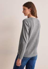 Harmaa pitkähihainen neule pehmeästä kankaasta, jossa on pyöreä pääntie ja ribbireunat olkapäillä, yhdistettynä sinisiin farkkuihin.