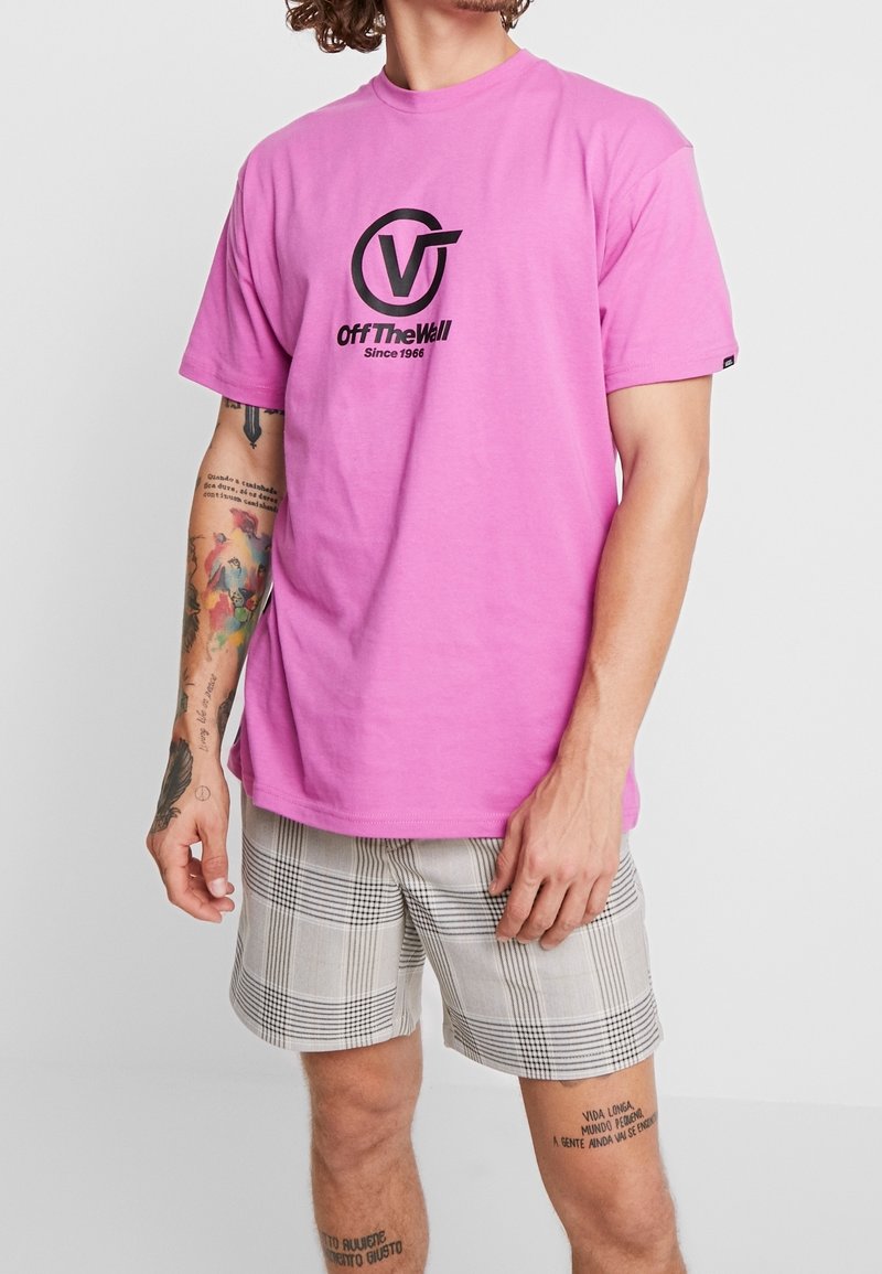 Vaaleanpunainen puuvillainen t-paita, jossa on musta pyöreä logo ja teksti "Off The Wall Since 1966" edessä, yhdistetty ruudullisiin shortseihin.