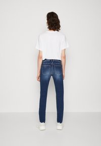Jeans de mezclilla azul oscuro con corte slim, cinco bolsillos y un leve desvanecimiento. Combinados con una camiseta blanca lisa y zapatillas blancas.