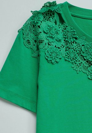 Top verde de manga corta con intrincada aplicación de encaje floral que decora el escote y la parte superior del hombro.