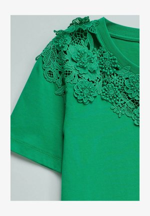 Haut vert à manches courtes avec une application en dentelle florale complexe décorant l'encolure et le haut de l'épaule.