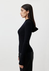 Čierny rebrovaný dlhý rukáv s priliehavým strihom a detailom kapucne. Textílie má hladkú štruktúru, nosený s elegantnými čiernymi nohavicami.