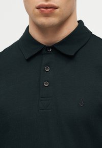 Άνδρας που φοράει σκούρο πράσινο πόλο πουκάμισο με τρία κουμπιά και μικρό κεντημένο λογότυπο στο αριστερό στήθος, εμφανίζεται από τον λαιμό μέχρι το επάνω μέρος του στήθους.