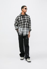 Μαύρο και γκρι καρό πουκάμισο oversized με κουμπιά μπροστά και τσέπη στο στήθος, συνδυασμένο με μαύρο παντελόνι με σκισίματα και διακοσμητικά καρφιά.