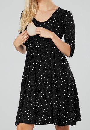 Žena v černých šatech s bílými puntíky, délky po kolena a s 3/4 rukávy, která odhrnuje část předního dílu, aby ukázala béžovou kojicí podprsenku.