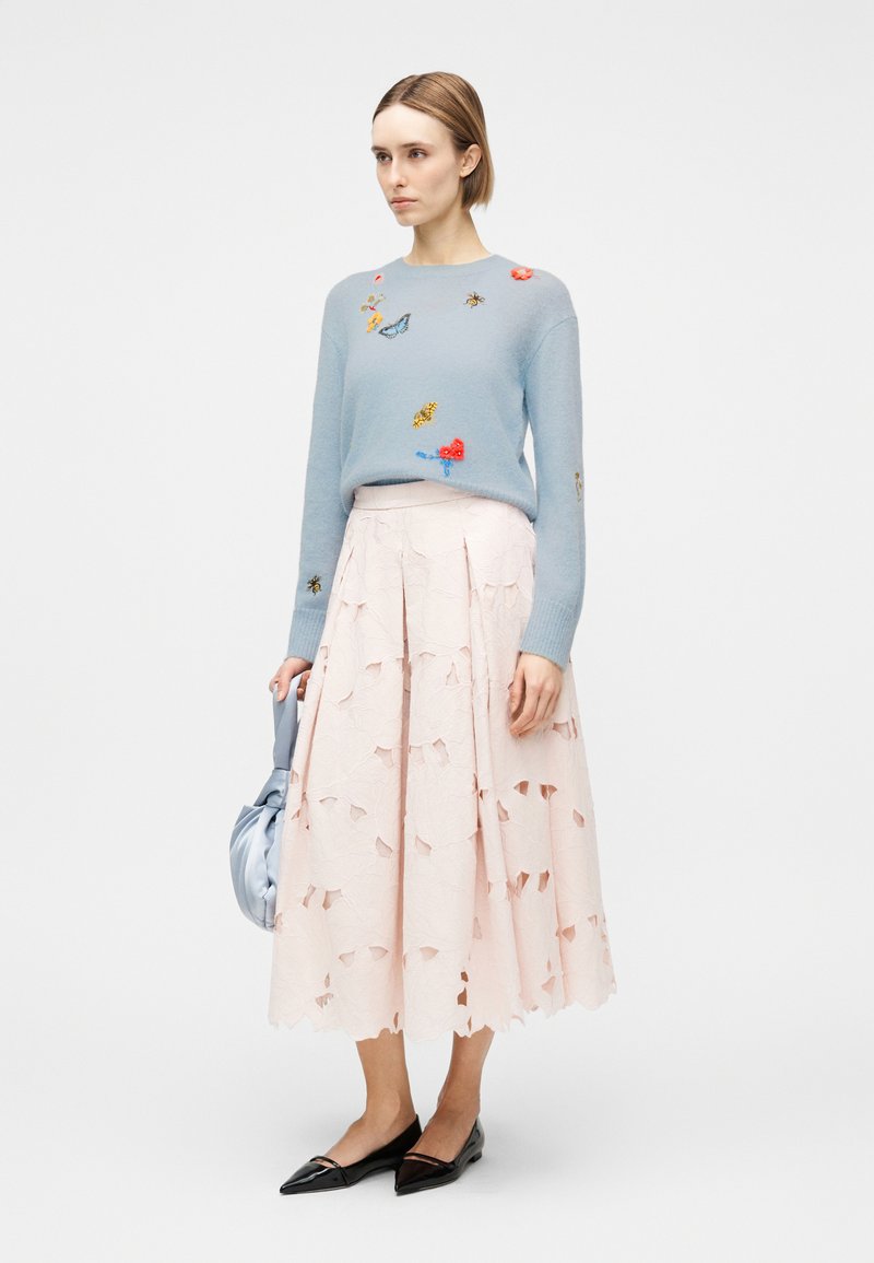 Donna che indossa un maglione azzurro con insetti e fiori ricamati, una gonna midi rosa pallido con ritagli, ballerine nere a punta, e tiene una borsa blu.