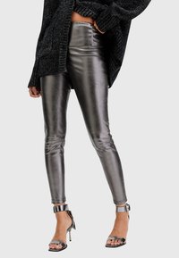Leggings metallici argento con una texture lucida, vestibilità slim e dettagli sui lati, abbinati a sandali neri con tacco a strappo.