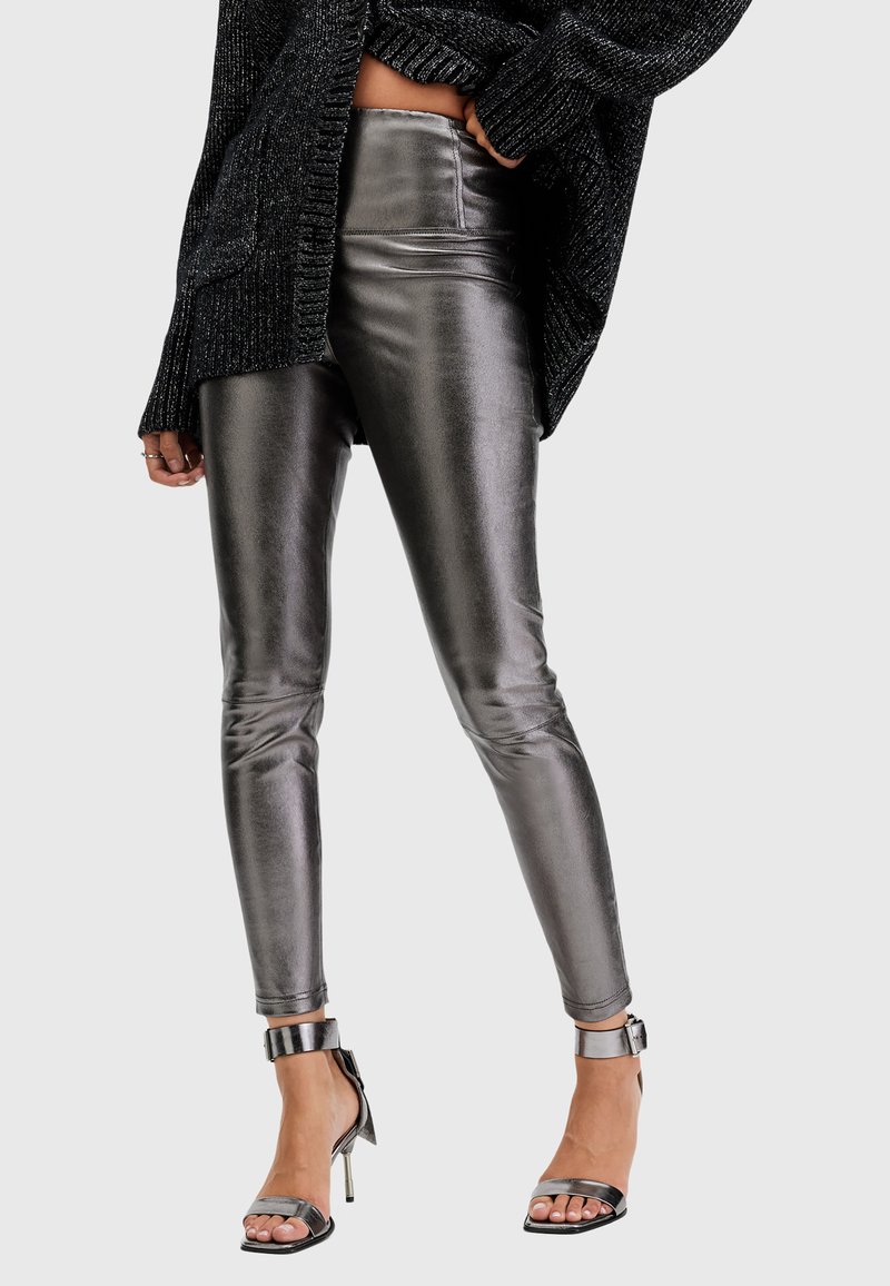 Leggings metallici argento con una texture lucida, vestibilità slim e dettagli sui lati, abbinati a sandali neri con tacco a strappo.