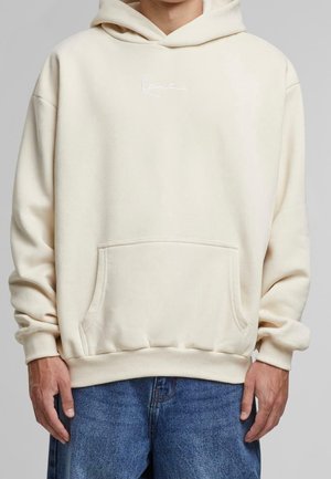 Person iført en lysebeige hættetrøje med forlomme og svag hvid tekst på brystet, kombineret med blå denimjeans.