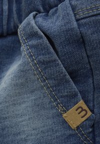 Denimtyg med en mörkblå nyans, med gula sömmar och en brun etikett med bokstaven "m." Närbild av materialets textur.