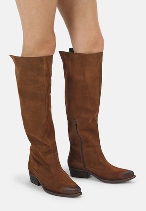 Hohe braune Wildlederstiefel mit niedrigen gestapelten Absätzen und Reißverschlüssen an der Innenseite, getragen von einer Person, die auf einem weißen Hintergrund steht.
