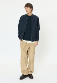 Jeune homme debout, les mains dans les poches, portant une veste zippée bleu marine, un pull foncé, un pantalon beige et des chaussures marron foncé, sur fond blanc.