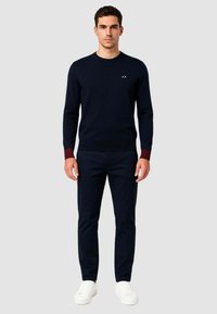 Maglione a maglia blu navy con polsini borgogna a contrasto, collo a giro e piccolo logo sul petto, abbinato a pantaloni scuri e sneaker bianche.