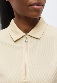 Κοντινό πλάνο ενός ατόμου που φοράει ένα μπεζ πόλο με υφή και ασημένιο φερμουάρ μερικώς κλειστό στον γιακά.