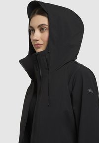 Black waterproof jacket with a high collar, adjustable hood, front zip, and logo patch on the sleeve. Smooth fabric texture.