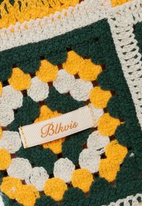 Tissu crocheté composé de carrés verts, jaunes et blancs, avec une étiquette en tissu blanc cousue brodée de "Blkvis" en fil jaune.