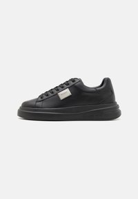 Zapatilla de cuero negro con punta redonda, suela plana y diseño de cordones. Presenta una placa de logo metálica en el lateral y una textura suave.