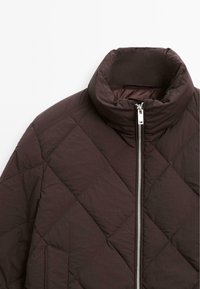Giacca trapuntata di colore marrone scuro con colletto alto e zip argentata. Presenta cuciture diagonali e una finitura in tessuto liscio e testurizzato.