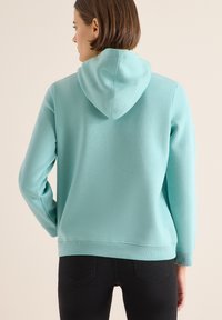 Felpa blu chiaro con cappuccio regolabile, realizzata in morbido materiale misto cotone. Presenta polsini e vita a costine. Indossata con pantaloni neri.
