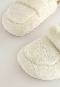 Pantofole di colore crema, morbide e pelose con una texture soffice e vellutata. Design con punta arrotondata e fascia larga sulla parte superiore. Nessun elemento metallico o accentuazioni visibili.