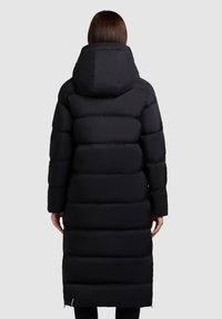 Persoon in een lange zwarte gewatteerde winterjas met een grote capuchon, gezien vanaf de achterkant tegen een effen achtergrond.