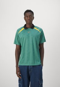 Polo-shirt vert à manches courtes avec un col noir et des accents jaunes, arborant un motif en relief subtil et un petit logo orange sur la poitrine.