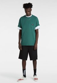 Camiseta verde de manga corta con cuello redondo y puños enrollados, con detalle de bolsillo, llevada sobre una camisa blanca de manga larga, combinada con pantalones cortos negros y calcetines con estampado.