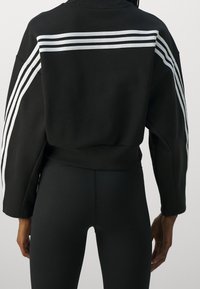 Felpa nera corta con vestibilit�à oversize. Presenta tre strisce bianche sulla schiena e polsini a coste. Indossata sopra leggings neri.