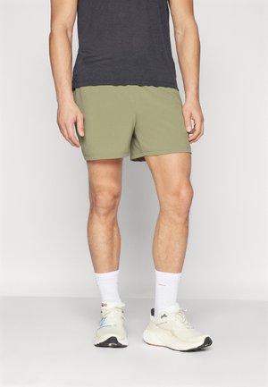 Uomo con pantaloncini verde oliva, calzini bianchi e scarpe da ginnastica bianche, in piedi contro uno sfondo chiaro e uniforme.