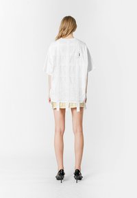 Camisa blanca de gran tamaño con un patrón de cuadros, que presenta tirantes verticales en la parte posterior. Combinada con una falda amarilla y zapatos de tacón negros.