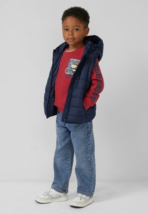 Kind in een marineblauwe hooded vest over een rode sweatshirt met grafisch ontwerp, blauwe jeans en witte sneakers, staand tegen een effen lichte achtergrond.