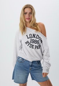 Svetlosivý oversized sveter s potlačou "LONDÝN 1986 HYDE PARK" v čiernej farbe. V kombinácii s modrými riflovými šortkami s roztrhnutým lemom.