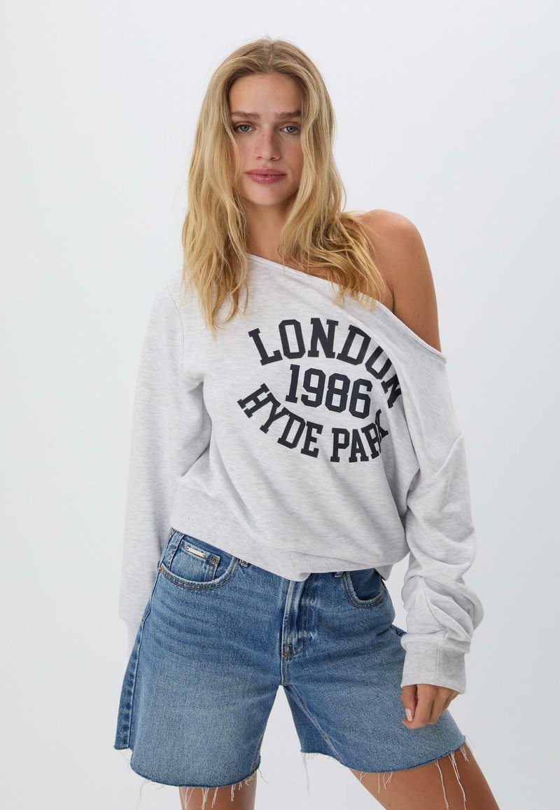 Svetlosivý oversized sveter s potlačou "LONDÝN 1986 HYDE PARK" v čiernej farbe. V kombinácii s modrými riflovými šortkami s roztrhnutým lemom.