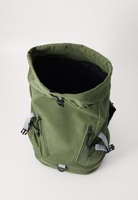 Zielony plecak typu roll-top z zamkami po bokach, regulowanymi ramiączkami i gładką teksturą. Wnętrze ma czarne podszycie.