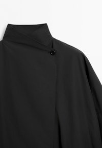 Černá bunda s vysokým límcem a asymetrickou přední klopou, zabezpečenou jedním černým knoflíkem na bílém pozadí.