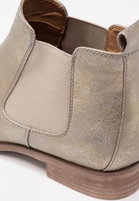 Gros plan d'une botte Chelsea en daim beige avec un panneau latéral élastique et un talon en bois empilé sur fond blanc.