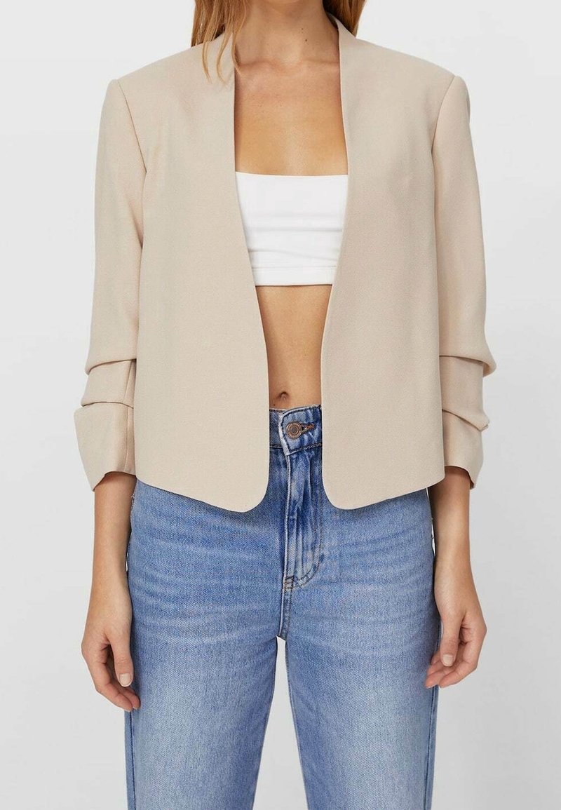 Blazer beige cropped con apertura frontale, maniche lunghe e una texture liscia, abbinato a un crop top bianco e jeans blu a vita alta.