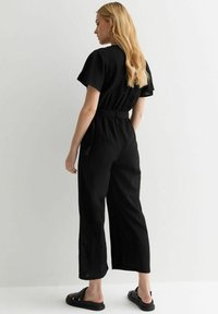 Jumpsuit noir à jambes larges, avec des manches courtes et une taille ceinturée. Le tissu semble léger et texturé, adapté à un usage décontracté.