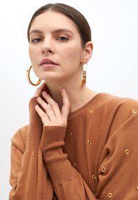 Bronzový svetr s metalickými detaily, dlouhé rukávy, žebrované manžety; velké zlaté kruhové náušnice; neutrální pozadí.