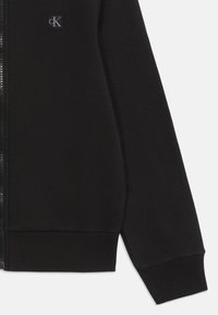 Čierna priedušná bunda na zips s rebrovanými manžetami a lemom, vyrobená z textúrovaného materiálu s malým vyšitým logom na ľavej hrudi.