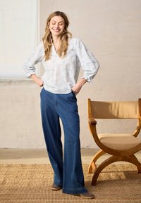 Femme souriante, les mains dans les poches, portant un chemisier blanc à motifs et un pantalon large bleu, debout à côté d'une chaise en bois dans une pièce aux tons neutres.