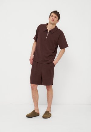 Muškarac u odgovarajućoj teksturiranoj smeđoj košulji kratkih rukava sa patentnim zatvaračem i kratkim hlačama stoji s jednom rukom u džepu na bijeloj pozadini.