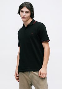 Young man with medium-length dark hair wearing a black polo shirt with red trim and beige pants, standing against a plain white background.