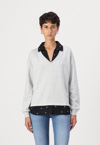 Felpa grigia a V con orlo in camicia a pois neri. Maniche lunghe e taglio rilassato, realizzata in un tessuto morbido con una texture liscia.