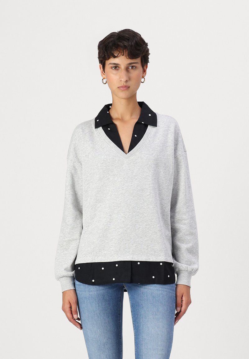 Felpa grigia a V con orlo in camicia a pois neri. Maniche lunghe e taglio rilassato, realizzata in un tessuto morbido con una texture liscia.
