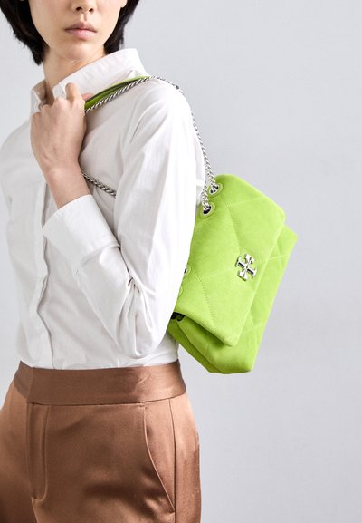 Borsa a tracolla verde in suede con design trapuntato, accessori in argento e catena, tenuta accanto a una persona che indossa una camicia bianca e pantaloni marroni.