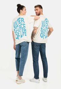 Lockere, cremefarbene Baumwoll-T-Shirts mit fettem blauen Text auf der Rückseite, der "WASSERQUELLE DES LEBENS" sagt. Getragen mit lässigen Jeans.