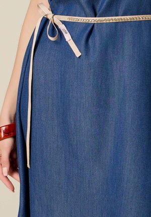 Taille d'une robe en denim bleu attachée avec une fine ceinture ruban beige comportant de petits strass et une étiquette de vêtement, main avec un bracelet marron visible.