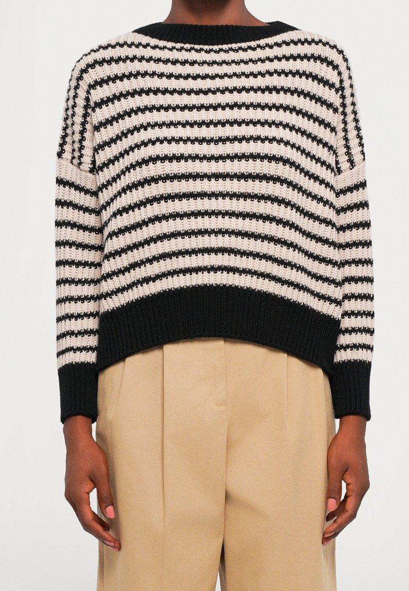 Pull en maille rayé noir et blanc avec bords côtelés porté avec un pantalon plissé beige sur une personne à la peau foncée.